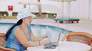 A woman in a sun hat sits at a table by the pool and works with a laptop remotely