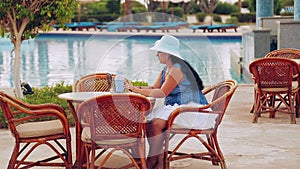 A woman in a sun hat sits at a table by the pool and works with a laptop remotely and drinks a cocktail