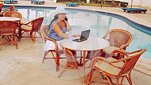 A woman in a sun hat sits at a table by the pool and works with a laptop remotely and speaks on the phone