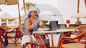 A woman in a sun hat sits at a table by the pool and works with a laptop remotely and speaks on the phone