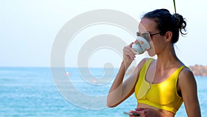 Woman in sunglasses drinks coffee in morning on ocean beach