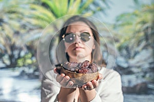 Woman in sunglasses holds out royal dates fruit in a bowl of coconut in a palm tree grove.