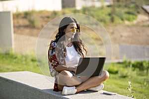 Woman in sunglasses looking away while using a laptop sitting on a bench in a park