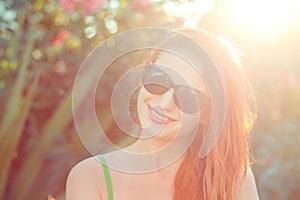 Woman with sunglasses smiling on a sunny day, sunflare on right on a green bush with pink flowers on background