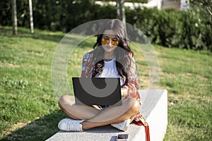 Woman with sunglasses using a laptop while sitting on a bench in a park