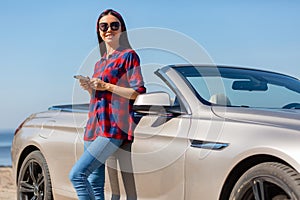 Woman in sunglasses using smartphone standing at the car and looking at the camera