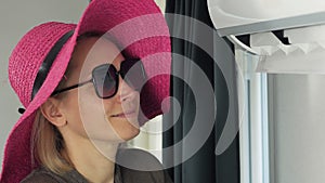 A woman with sunglasses and a wide-brimmed hat is standing near the air conditioner enjoying the coolness.