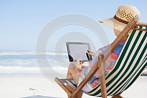 Woman in sunhat sitting on beach in deck chair using tablet pc