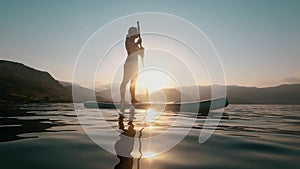 Woman on a SUP board paddling through shining water at sunrise.