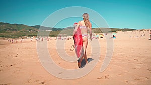 Woman surfer in bikini walking with red surfboard on sandy beach