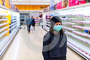 Woman with surgical mask looks confused in the aisle between empty shelves in a supermarket