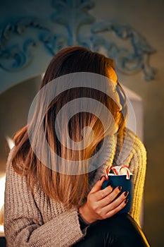 Woman in sweater with a cup of cocoa by the fireplace.