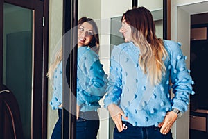 Woman in sweater looking at mirror. Girl trying clothes on by wardrobe at home. Beauty fashion