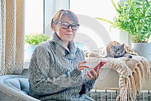 Woman in sweater sitting in armchair near window with heating radiator and sleeping cat