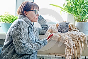 Woman in sweater sitting in armchair near window with heating radiator and sleeping cat