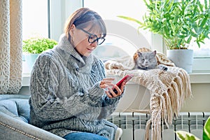 Woman in sweater sitting in armchair near window with heating radiator and sleeping cat