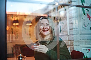 Woman in sweater sitting in restaurant with cup of fruit tea and looking out the window  view through glass