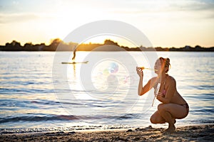 Woman in a swimsuit blowing soap bubbles walking along the beach near the lake on summer vacation