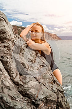 Woman swimsuit sea. Attractive blonde woman in a black swimsuit enjoying the sea air on the seashore around the rocks