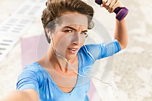 Woman in swimsuit sitting on yoga mat looking in camera with dumbbell in hand and taking selfie