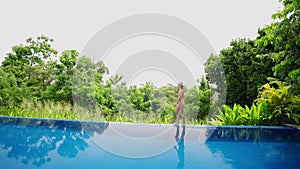 Woman in swimsuit strolls by luxurious villa infinity pool, green tropical landscape. Side view of leisure walk