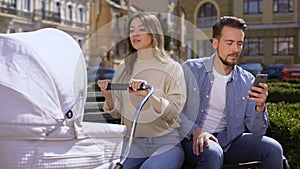 Woman swinging newborn carriage looking angrily at husband using smartphone