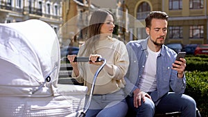 Woman swinging newborn carriage looking angrily at husband using smartphone