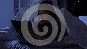 woman with tablet pc under blanket in bed at night