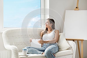 Woman with Tablet Thinking Looking forward at Home Sofa. Beautiful Girl Dreaming with Touch Pad next to Window and Drawing Easel