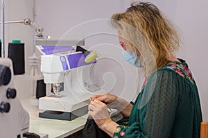 Woman tailor with a medical mask on her face works in a sewing atelier, work during quarantine