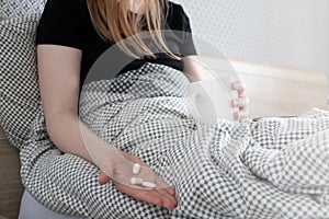 Woman take pills. Antibiotic or painkiller in hand with selective focus and white cup at bed. Woman feeling sick at home, indoors