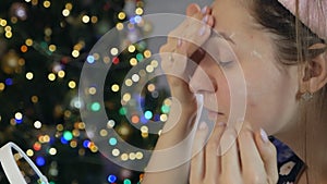 A woman takes care of her beautiful skin on her face, standing near the Christmas tree. Young woman applying moisturizer