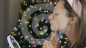 A woman takes care of her beautiful skin on her face, standing near the Christmas tree. Young woman applying moisturizer