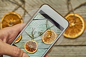 Woman takes a photo of christmas decoration made of dried orange slices on her white modern smarthfone. Taking blogging conent for