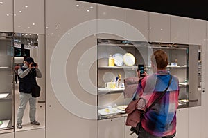 A woman takes photographs of exhibits in an exhibition hall with her smartphone. Photographer man reflected in the mirror