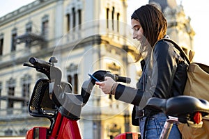 Woman taking a bike with her cell phone in a public parking of rental bicycles