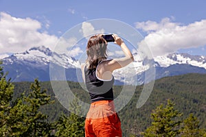 Woman taking cell phone photo of majestic mountain range, snow covered peaks, Canadian nature, Tantulus Range, tourist.