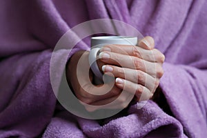 Woman taking cover under lilac plush blanket and her hands holding and warming on white ceramic cup of hot drink. Cosiness and slo