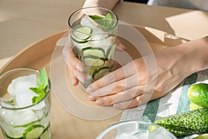 Woman taking glass of fresh cucumber water from wooden tray
