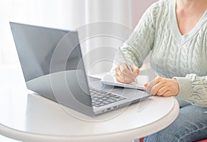 Woman taking notes in notebook while using laptop with at home. freelancer writing details on book while working on laptop in