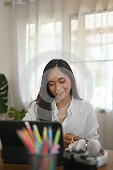 A woman is taking notes while sitting in front of a computer tablet at the wooden working desk