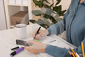 Woman taking notes while using smartphone at white marble table indoors, closeup