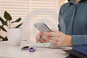 Woman taking notes while using smartphone at white marble table indoors, closeup