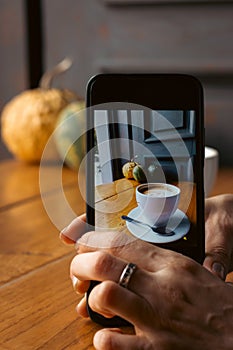 Woman taking a photo of morning coffee. Coffee cup with pumpkins on background in phone screen. Blogging concept.