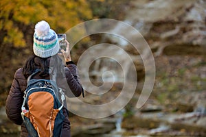 woman taking a photo of a waterfall in autumn with changing leaves on her mobile phone from behind