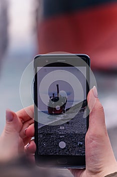 Woman taking pictures of a large ship on her cell phone while walking along the beach