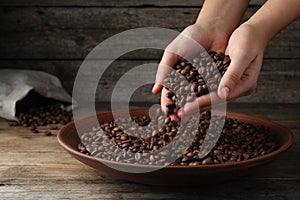 Woman taking pile of roasted coffee beans from bowl at wooden table, closeup