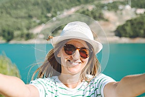 Woman taking selfie photo with wide angle front camera of mountains lake background. Traveler female having fun on the