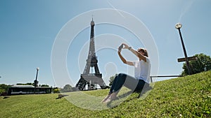 Woman is taking selfie using smartphone sitting near Eiffel tower in Paris in daytime,