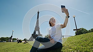 Woman is taking selfie using smartphone sitting near Eiffel tower in Paris in daytime, Wide footage